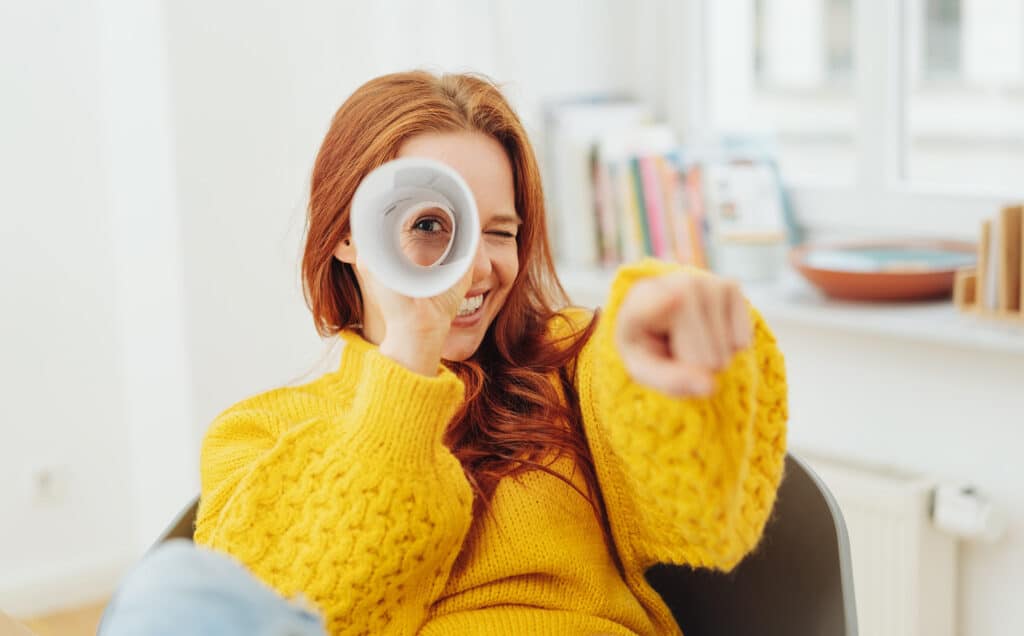Woman pointing at camera while looking through roll of paper as if it were a spyglass