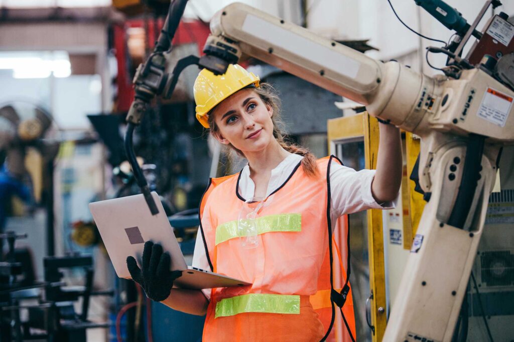 Happy female industrial engineer or technician worker in hard helmet and uniform using laptop checking on robotic arm machine.