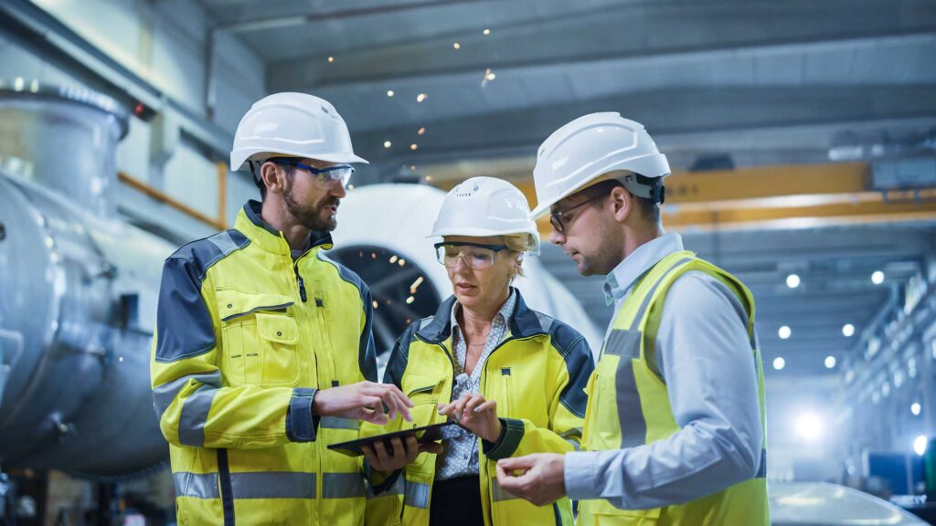 Three Heavy Industry Engineers Stand in Pipe Manufacturing Factory, Use Digital Tablet Computer, Have Discussion. Large Pipe Assembly.