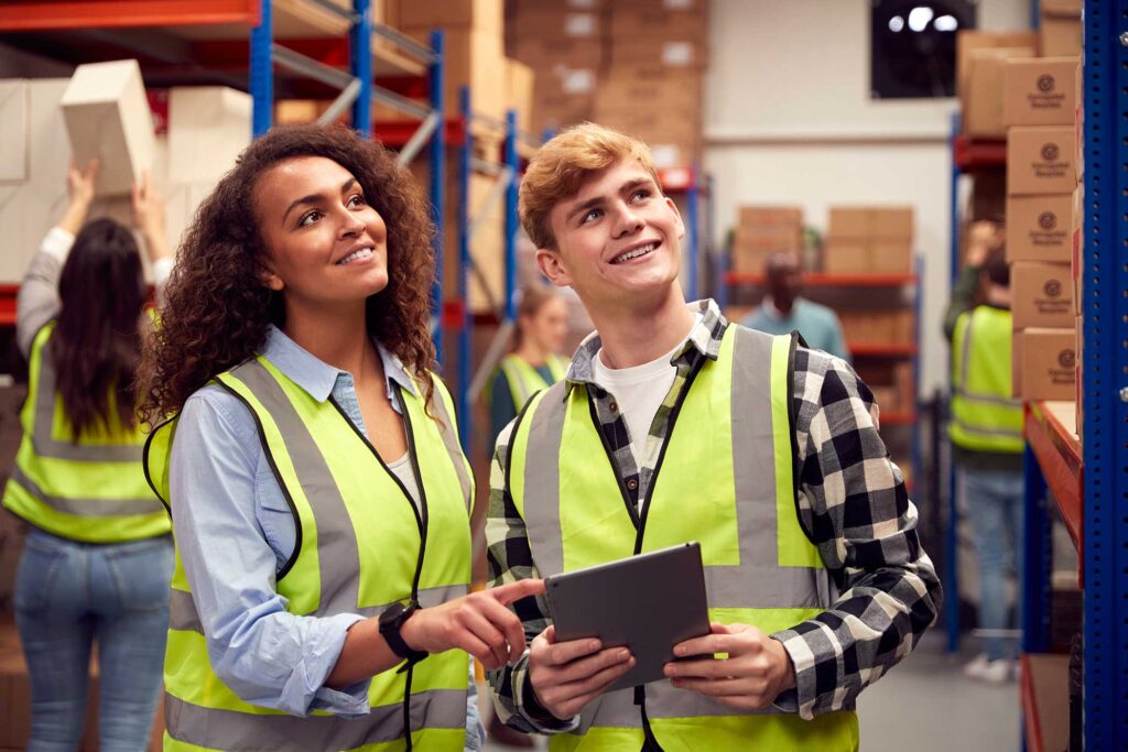 Male Intern With Team Leader Looking At Digital Tablet Inside Busy Warehouse Facility