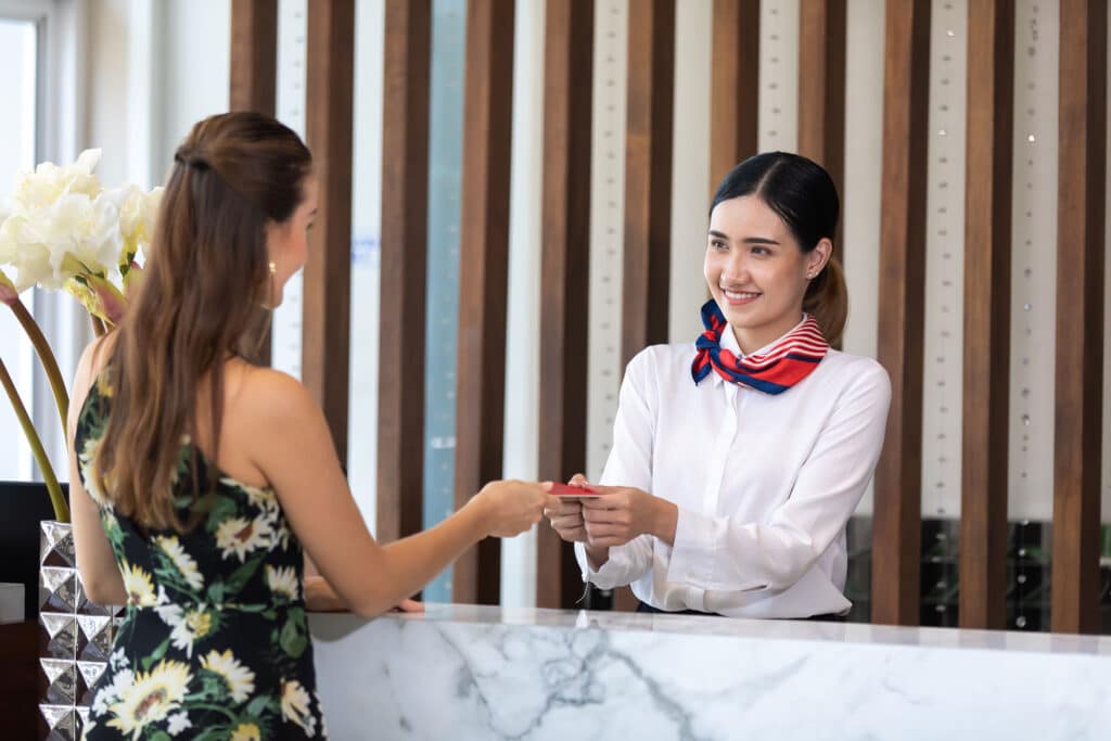 Smiling young beautiful Asian female receptionist giving key card to customer hand at hotel reception counter desk